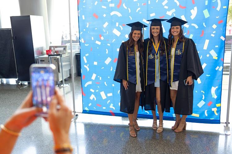 Three graduating students pose together in front of a backdrop as someone takes a photo of them on a phone